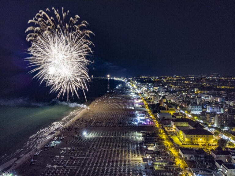 Fuochi d'artificio sulla spiaggia illuminata di notte, vista aerea di un lungomare urbano.