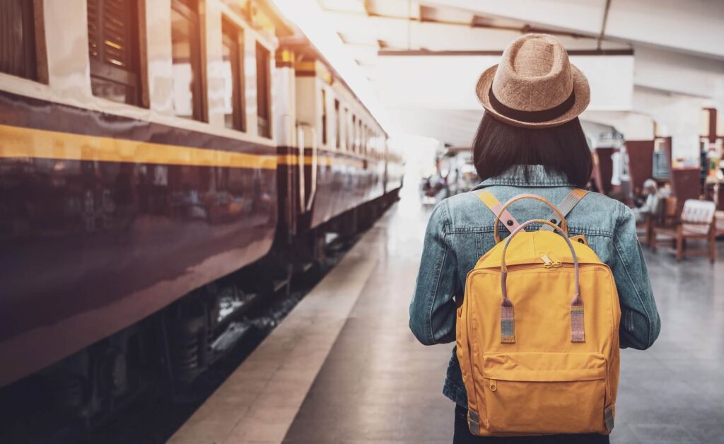 Eine Person mit Hut und gelbem Rucksack steht an einem Bahnhof neben einem Zug.