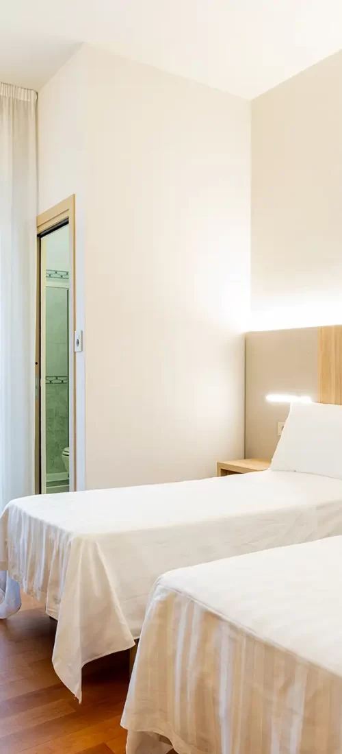 A bright hotel room with two single beds, wooden furniture, and a window with sheer curtains.