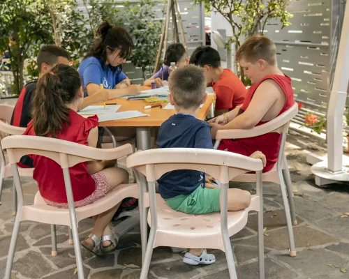 Children and a teacher sit around a table outdoors, writing and drawing.