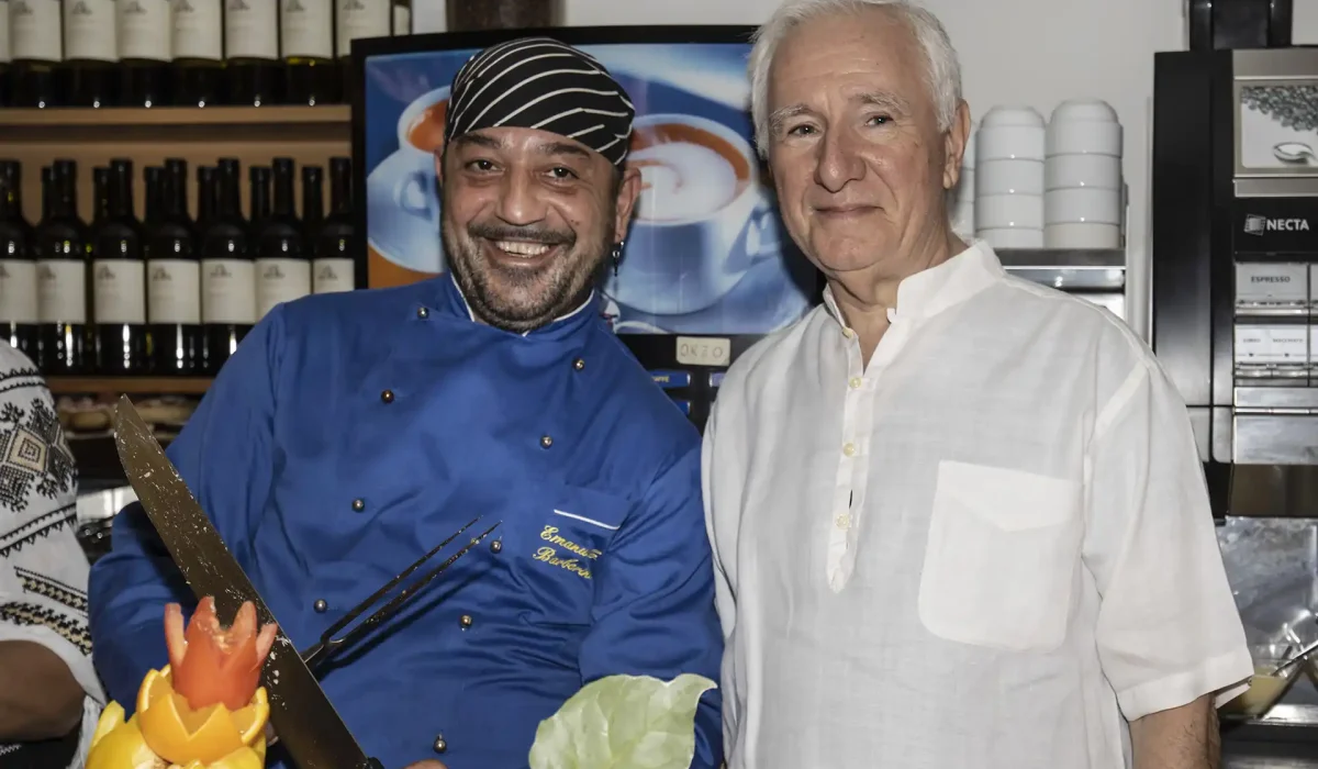 Two smiling men, one in a blue chef's jacket and striped headscarf, stand together with culinary decorations in the foreground.