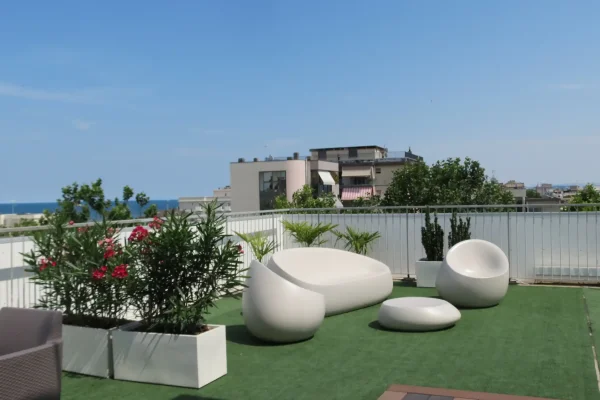 A rooftop patio with modern white furniture, planters, and a view of buildings and the sea.