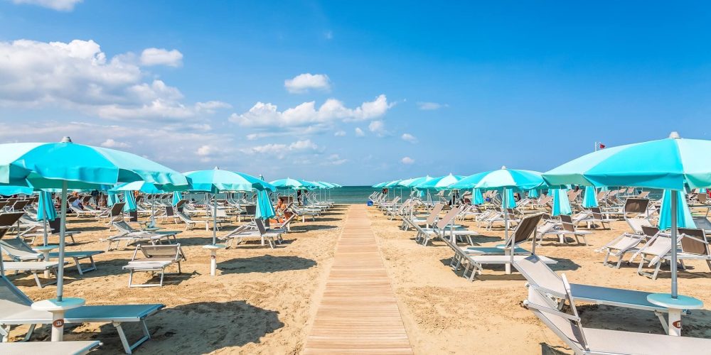 Spiaggia sabbiosa con ombrelloni azzurri e lettini allineati sotto un cielo sereno.